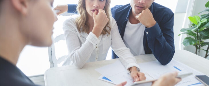 Real estate agent with couple looking through documents. The agent is holding a digital tablet showing it to the clients. Couple are casually dressed. They are looking concerned and worried and upset. Over the shoulder view.